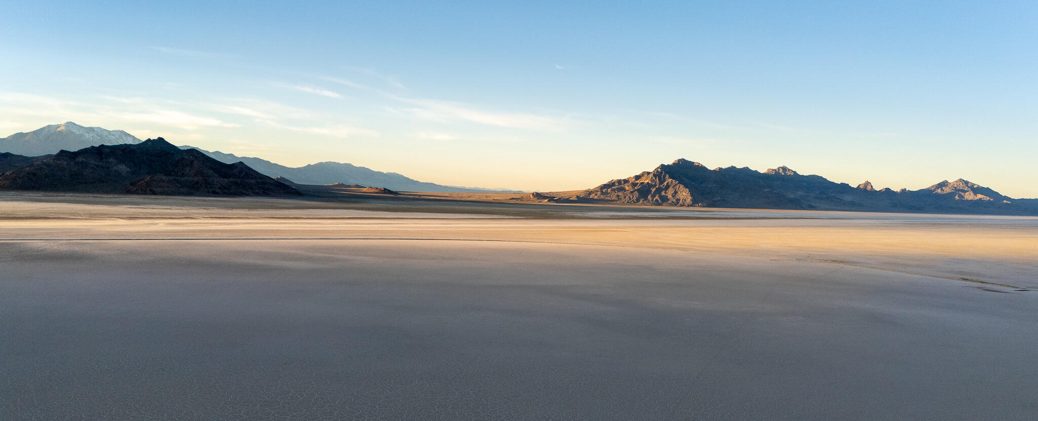 Wide aerial view of a vast salt flat with soft ripples, mountains in the distance, and pale blue sky at sunrise.